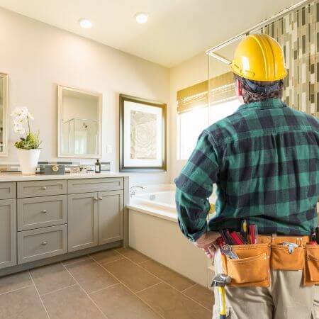 A bathroom remodeling professional in a hard hat standing in a bathroom, inspecting the construction work.