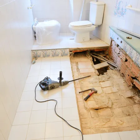 A bathroom during floor demolition with a jackhammer and exposed pipes.